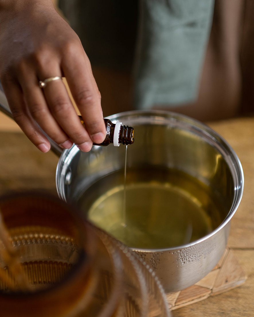 Žena nalieva esenciálny olej do hrnca s olejom. Používa pri tom pomôcky na výrobu kozmetiky. Nachádza sa v tvorivej dielničke. 